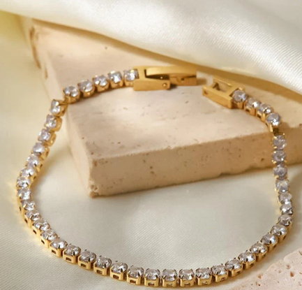 A gold tennis bracelet with sparkling stones displayed on a beige cushion.