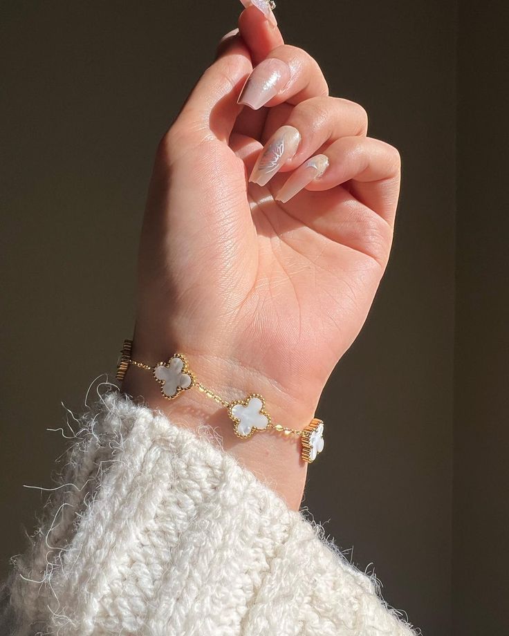 Hand wearing a gold bracelet with clover charms against a neutral background